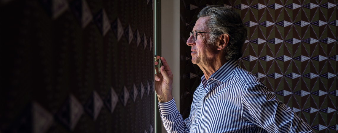 Mann im gestreiften Hemd blickt aus einem Fenster, umgeben von gemusterten Wänden.