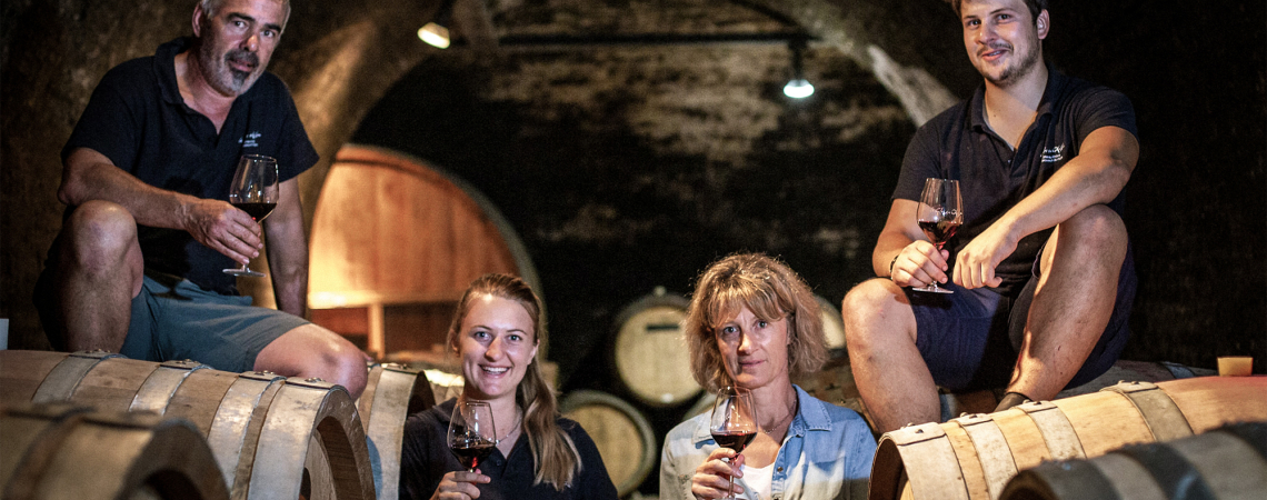 Vier Personen sitzen auf Weinfässern in einem atmosphärischen Weinkeller mit Weingläsern.