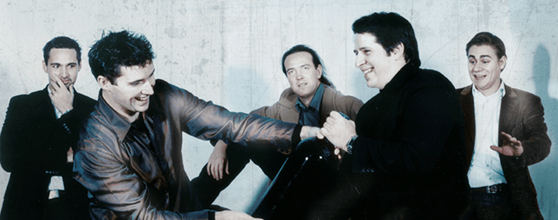 Fünf Männer in Anzügen lachen und kämpfen spielerisch um eine große Flasche.