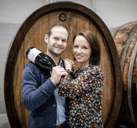 Zwei lächelnde Personen vor Weinbarrique, halten eine Weinflasche.