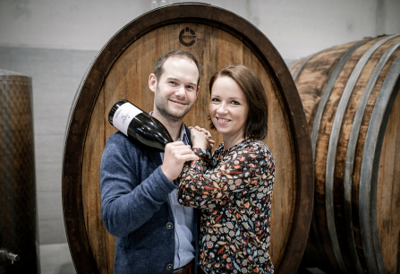 Zwei lächelnde Personen vor Weinbarrique, halten eine Weinflasche.