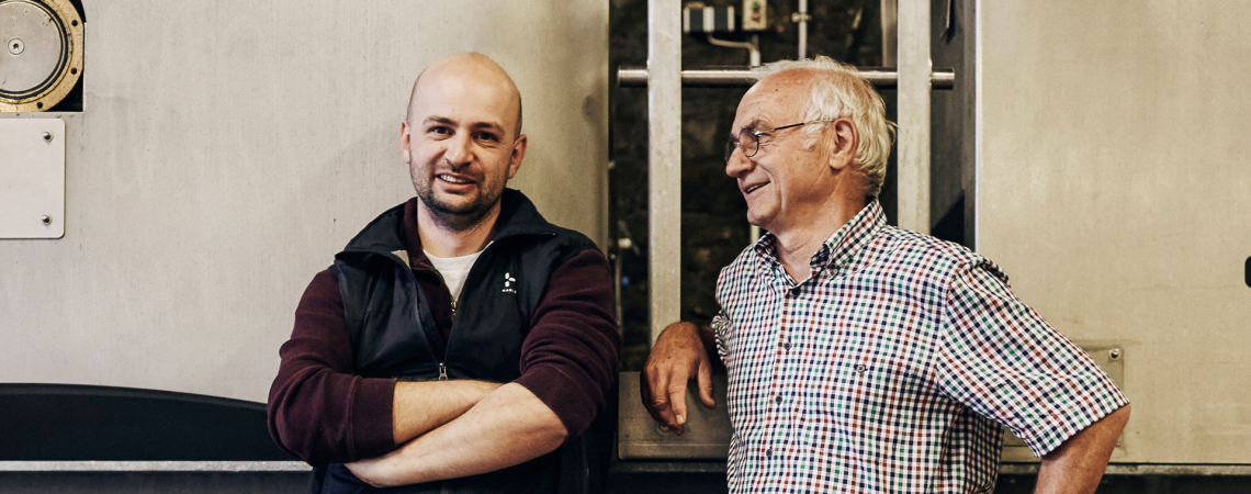 Zwei lächelnde Männer in Arbeitskleidung vor einer Industriemaschine.