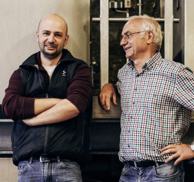 Zwei lächelnde Männer in Arbeitskleidung vor einer Industriemaschine.