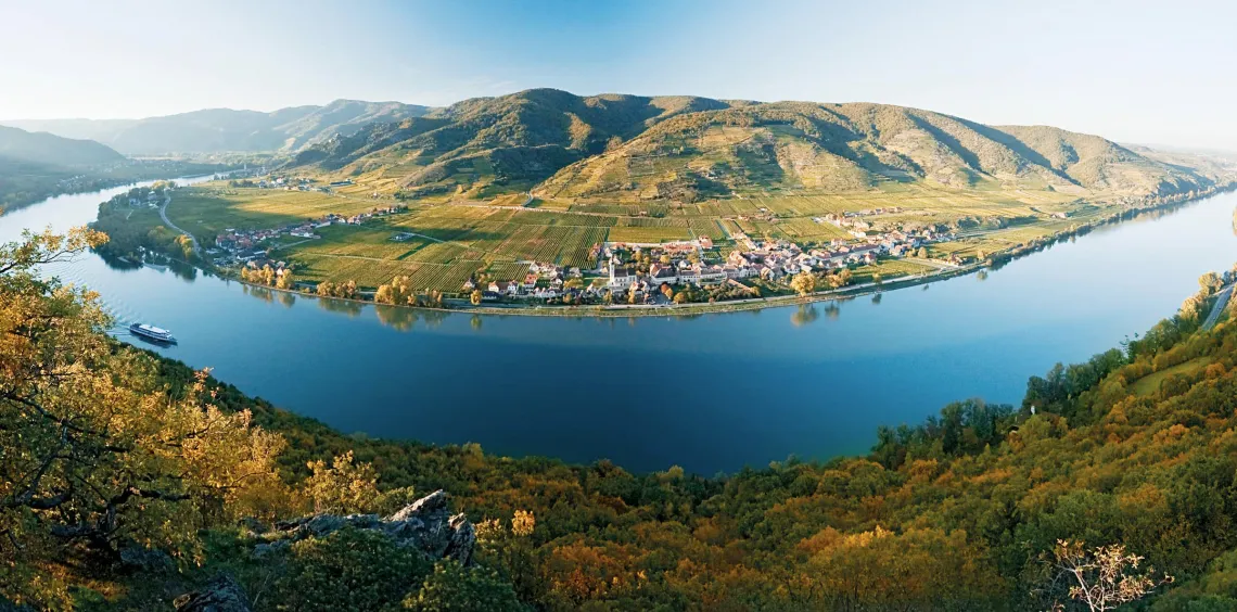 Landschaft mit Fluss, umliegenden Hügeln und malerischem Dorf bei klarem Himmel.