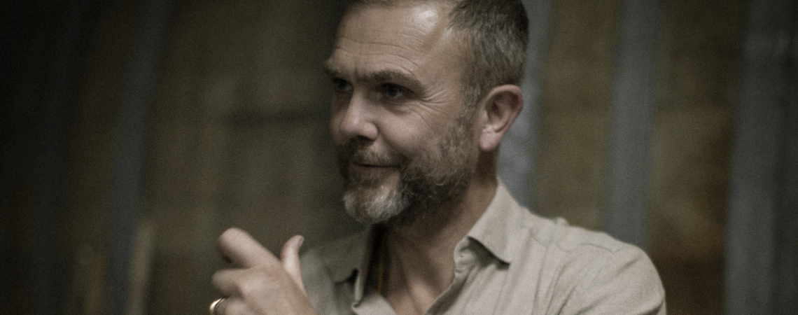 Mann mit Bart und Hemd, lächelnd, vor unscharfem Hintergrund.