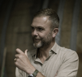 Mann mit Bart und Hemd, lächelnd, vor unscharfem Hintergrund.