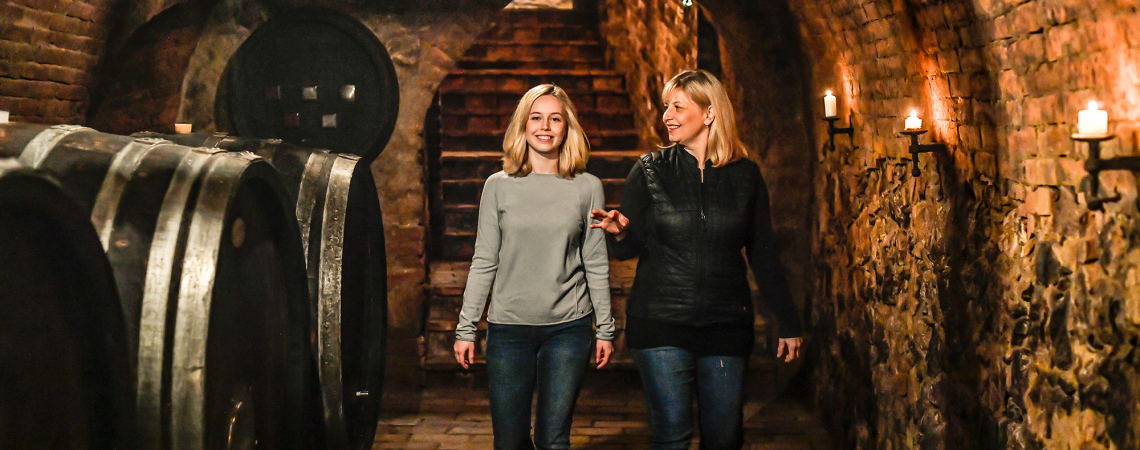 Zwei Personen gehen lächelnd durch einen historischen Weinkeller mit brennenden Kerzen.