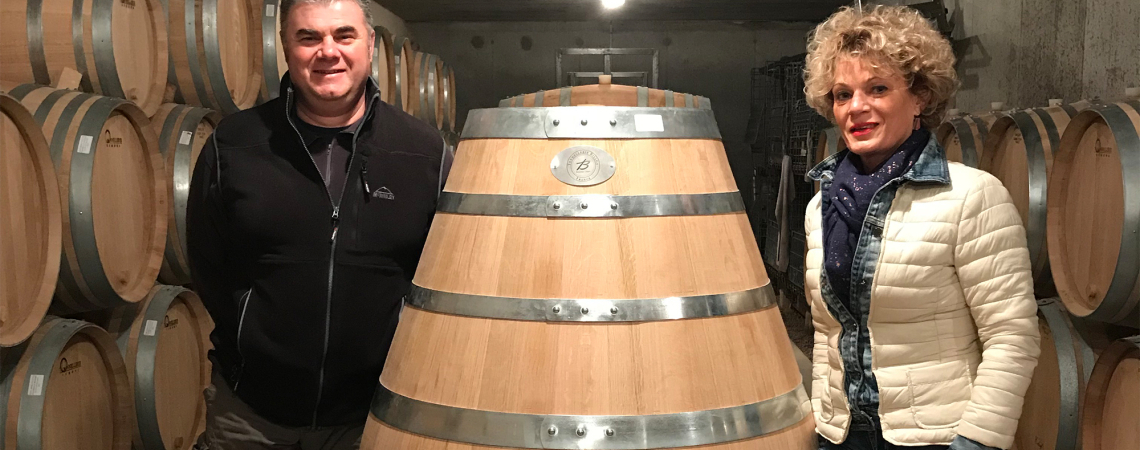 Mann und Frau stehen in Weinkeller neben großem Holzfass, umgeben von Fässern.