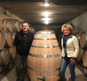 Mann und Frau stehen in Weinkeller neben großem Holzfass, umgeben von Fässern.