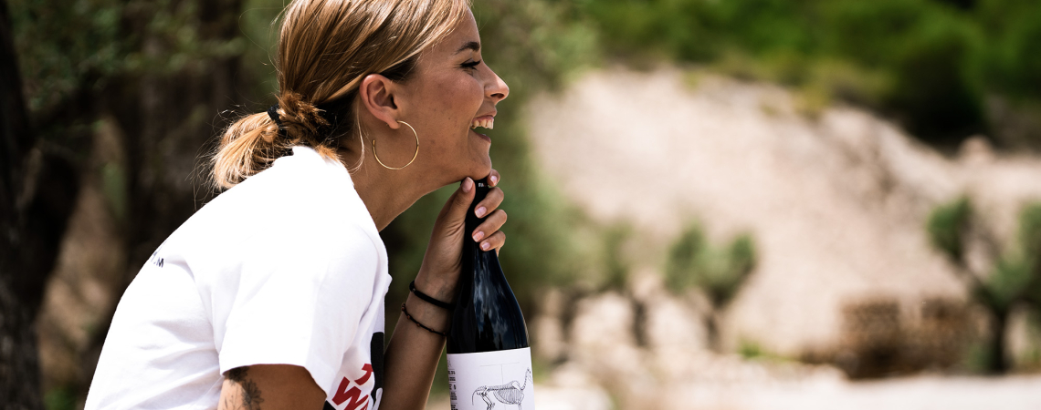 Frau im Freien lacht und hält eine Weinflasche, umgeben von Bäumen.