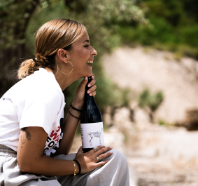 Frau im Freien lacht und hält eine Weinflasche, umgeben von Bäumen.