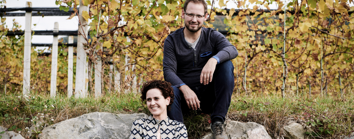 Zwei Personen sitzen auf Steinen vor herbstlichen Weinreben.