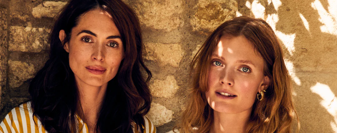 Zwei Frauen in sommerlicher Kleidung halten Champagnergläser vor einer Steinwand.