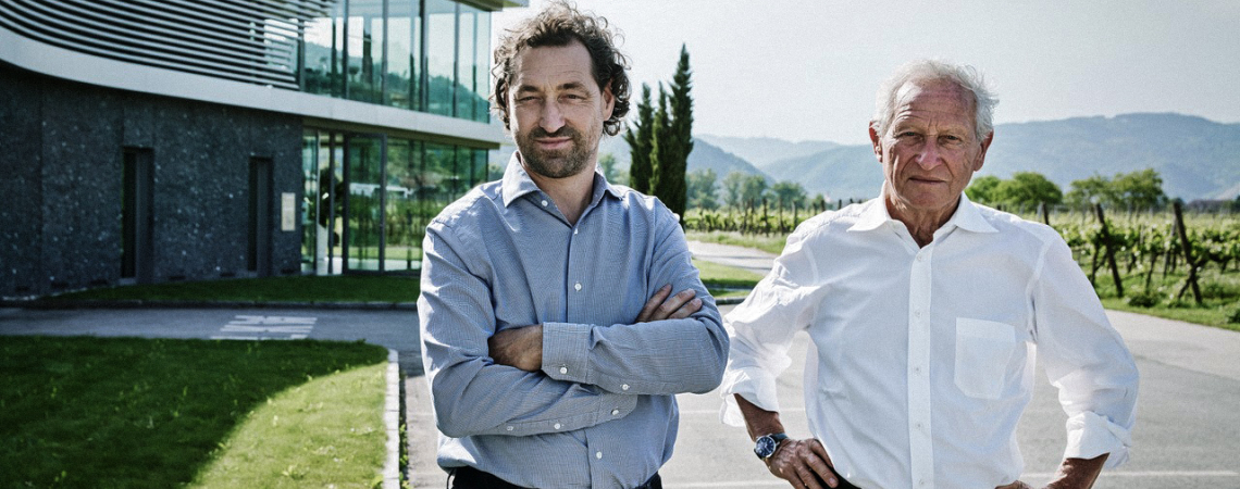 Zwei Männer vor einem modernen Gebäude in einer Weinberglandschaft.