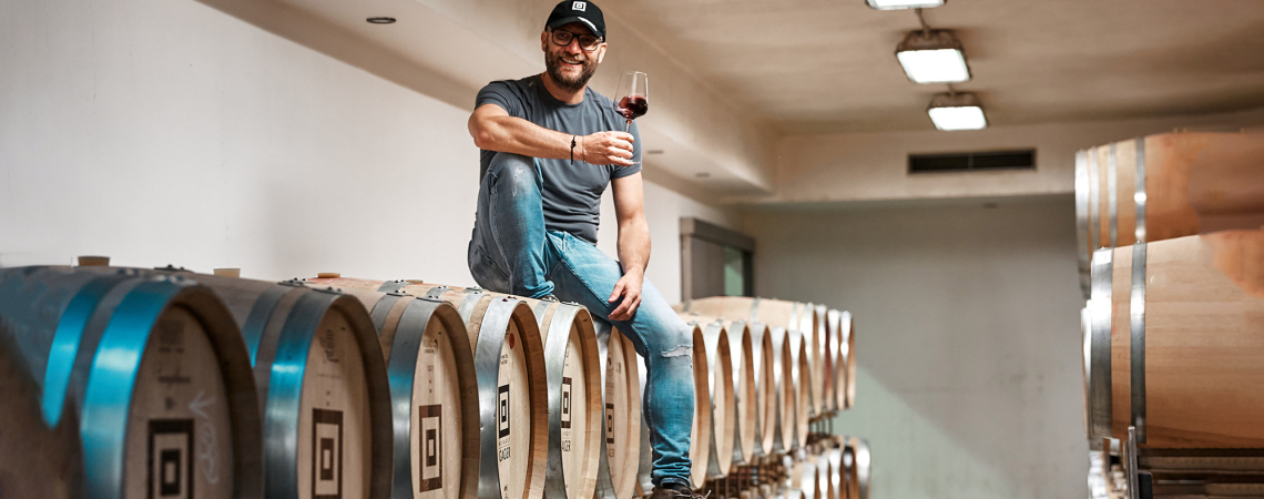 Mann mit Weinglas sitzt auf Holzfässern in einem Keller.