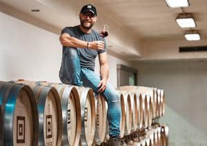 Mann mit Weinglas sitzt auf Holzfässern in einem Keller.