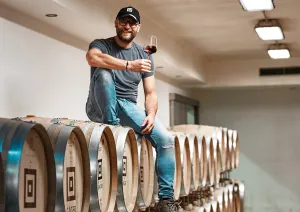 Mann mit Weinglas sitzt auf Holzfässern in einem Keller.