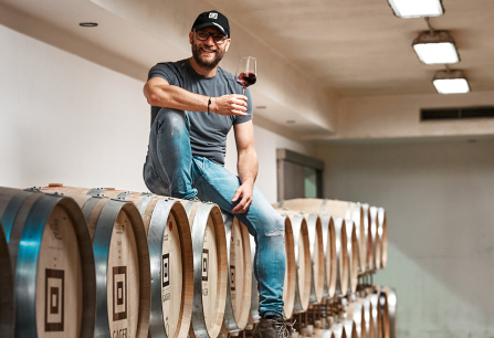 Mann mit Weinglas sitzt auf Holzfässern in einem Keller.