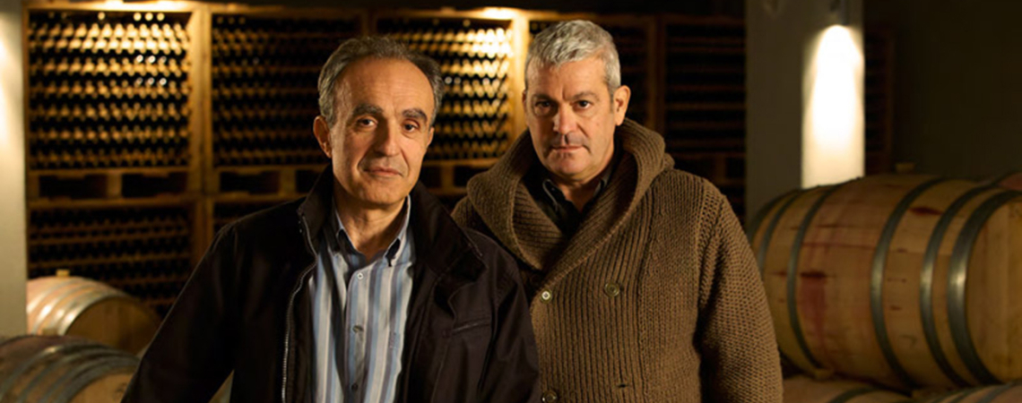 Zwei Männer stehen vor Weinfässern in einem stimmungsvoll beleuchteten Weinkeller.