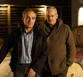 Zwei Männer stehen vor Weinfässern in einem stimmungsvoll beleuchteten Weinkeller.