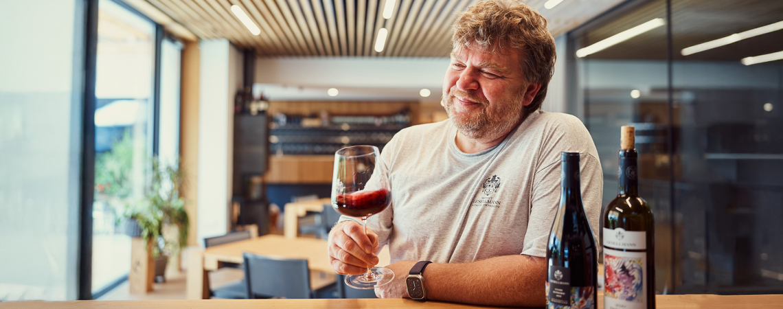 Mann verkostet Wein in modernem, holzverkleidetem Raum mit Flaschen auf dem Tisch.