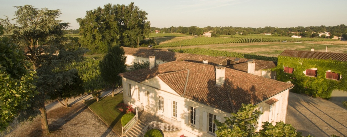 Helles Landhaus mit Ziegeldach inmitten grüner Landschaft, umgeben von Bäumen.