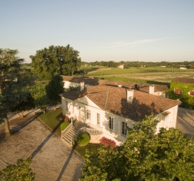 Helles Landhaus mit Ziegeldach inmitten grüner Landschaft, umgeben von Bäumen.