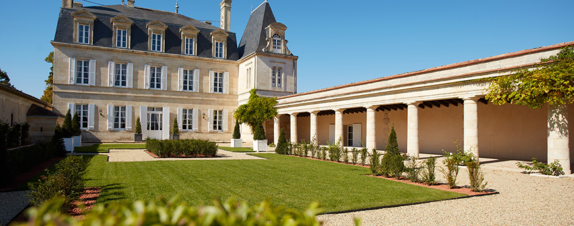 Historisches Herrenhaus mit gepflegtem Garten, flankiert von einer langen Arkade unter blauem Himmel.