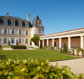 Historisches Herrenhaus mit gepflegtem Garten, flankiert von einer langen Arkade unter blauem Himmel.