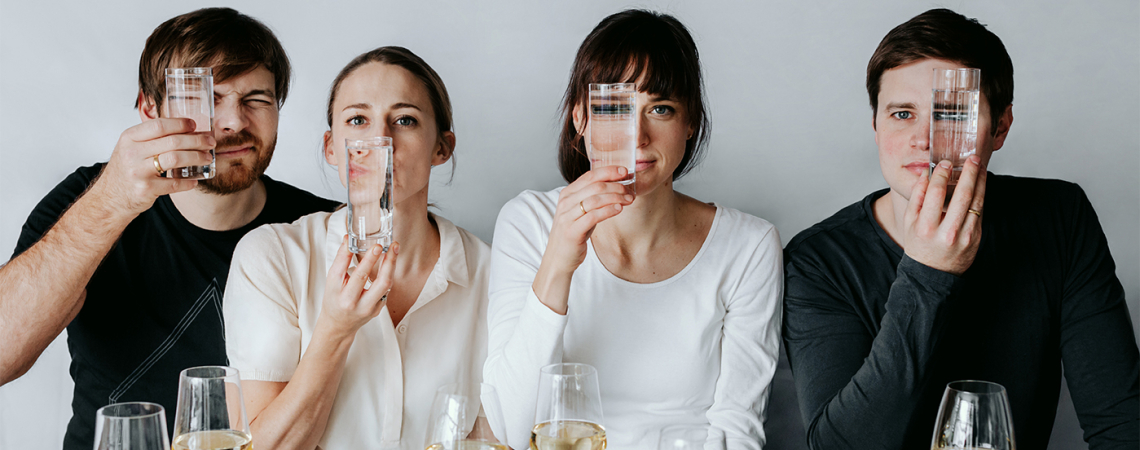 Vier Personen halten Gläser an ihre Gesichter hinter gedecktem Tisch.