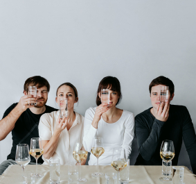 Vier Personen halten Gläser an ihre Gesichter hinter gedecktem Tisch.