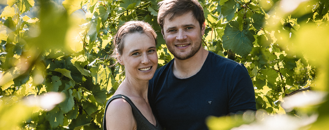 Paar steht lächelnd zwischen üppigen grünen Weinreben im Sonnenlicht.