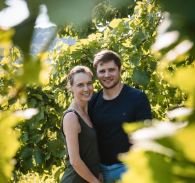 Paar steht lächelnd zwischen üppigen grünen Weinreben im Sonnenlicht.