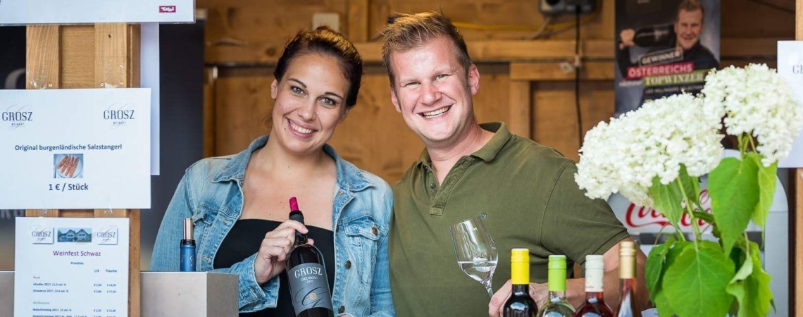 Zwei lächelnde Personen präsentieren Weinflaschen an einem rustikalen Verkaufsstand.