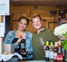 Zwei lächelnde Personen präsentieren Weinflaschen an einem rustikalen Verkaufsstand.