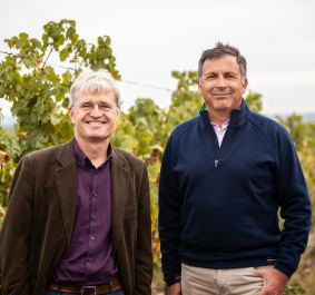 Zwei Männer lächeln vor grünen Weinreben im Freien.