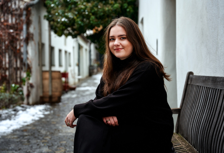 Frau in schwarzer Kleidung sitzt lächelnd auf einer Bank in einer Gasse.