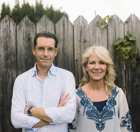 Zwei Personen vor Holzzaun, lächelnd, in Jeans und verzierten Oberteilen.