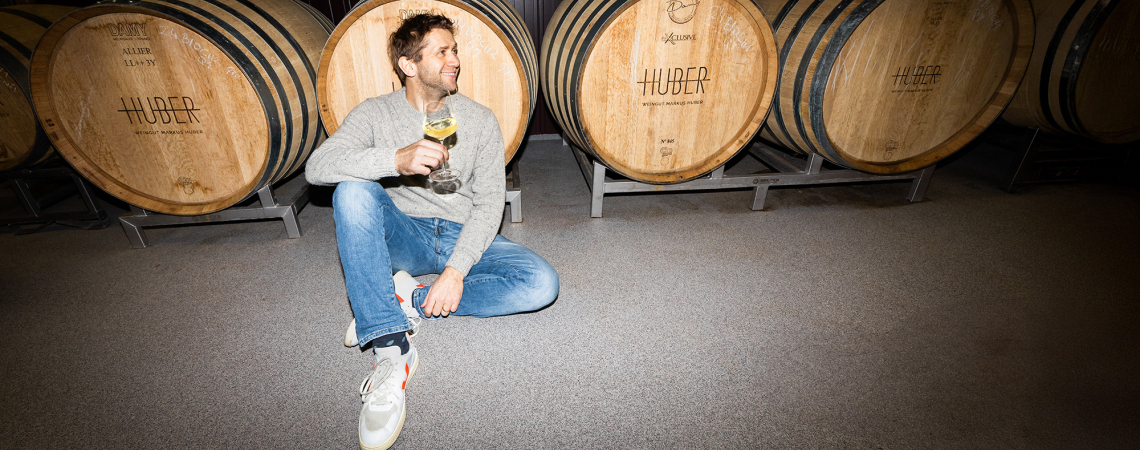 Mann in Jeans vor Weinfässern sitzt entspannt mit Getränk in der Hand.