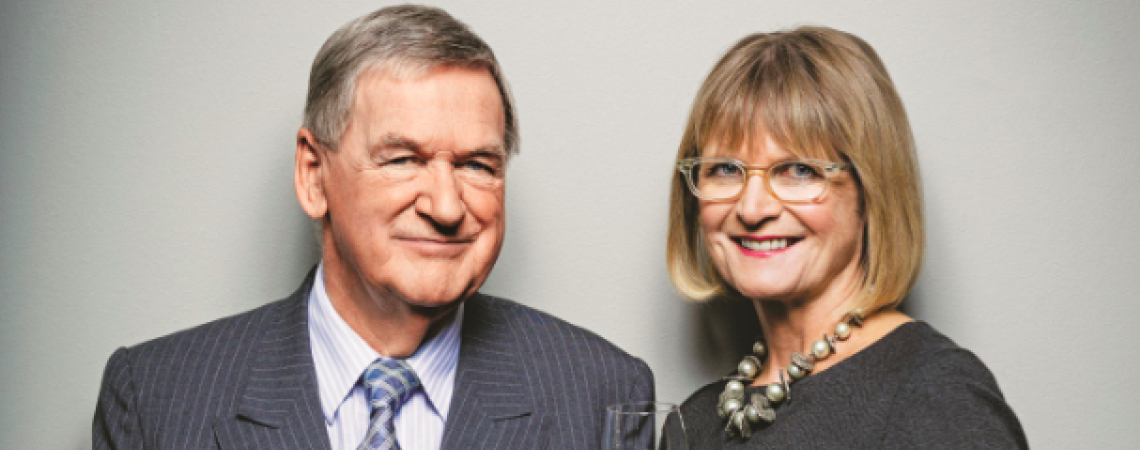 Elegantes Paar im Anzug und Kleid hält Weingläser vor neutralem Hintergrund.