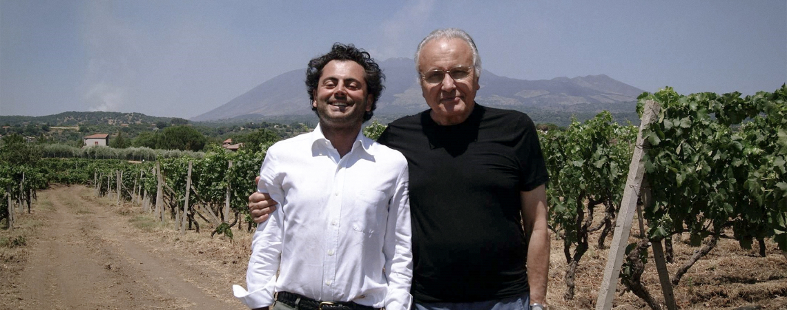 Zwei Männer in einem sonnigen Weinberg vor einem Berg im Hintergrund.