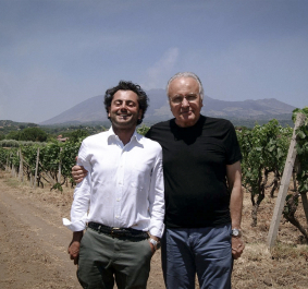 Zwei Männer in einem sonnigen Weinberg vor einem Berg im Hintergrund.