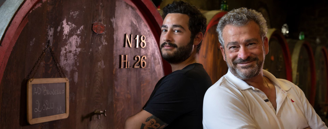 Zwei lächelnde Männer stehen vor großen Holzfässern in einem Weinkeller.