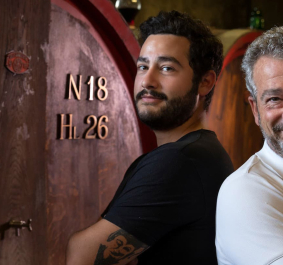 Zwei lächelnde Männer stehen vor großen Holzfässern in einem Weinkeller.