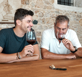 Zwei Männer genießen Rotwein in einem rustikalen Raum mit Steinfassade und Holztisch.