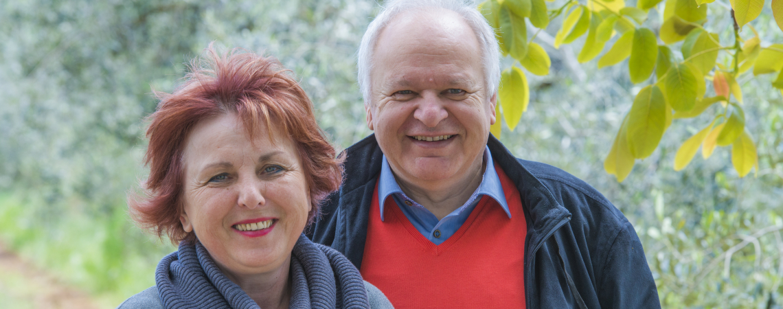Älteres Paar lächelt im Grünen mit herbstlichem Laub im Hintergrund.