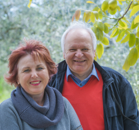 Älteres Paar lächelt im Grünen mit herbstlichem Laub im Hintergrund.