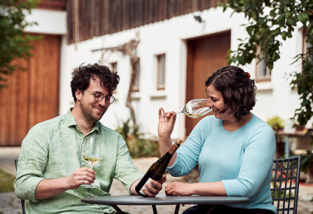 Zwei Personen genießen Weißwein an einem Tisch im Freien vor traditionellem Gebäude.