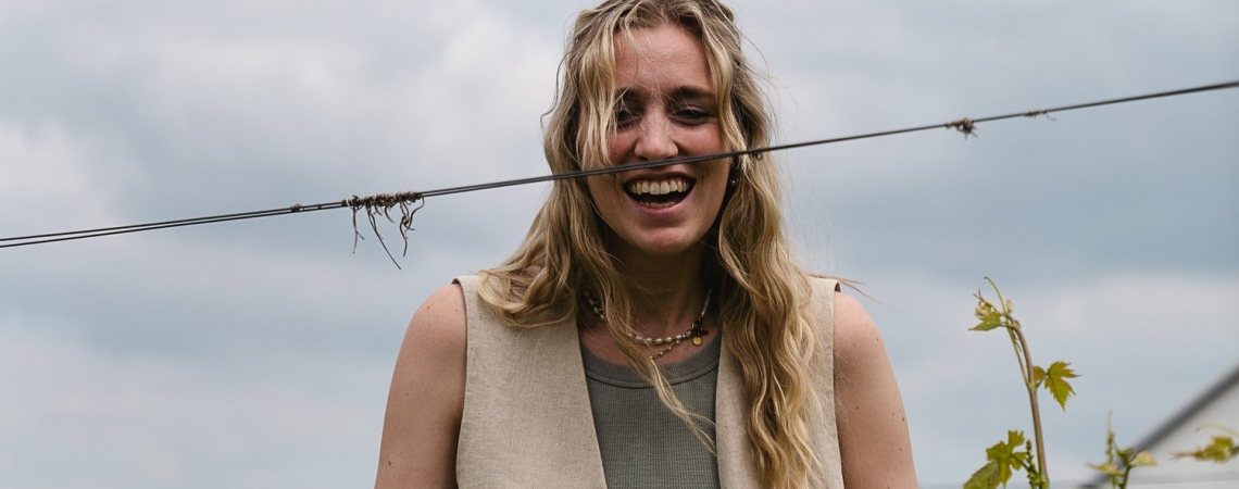 Frau in beigener Weste steht lächelnd im Weinberg, bewölkter Himmel.