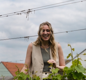 Frau in beigener Weste steht lächelnd im Weinberg, bewölkter Himmel.